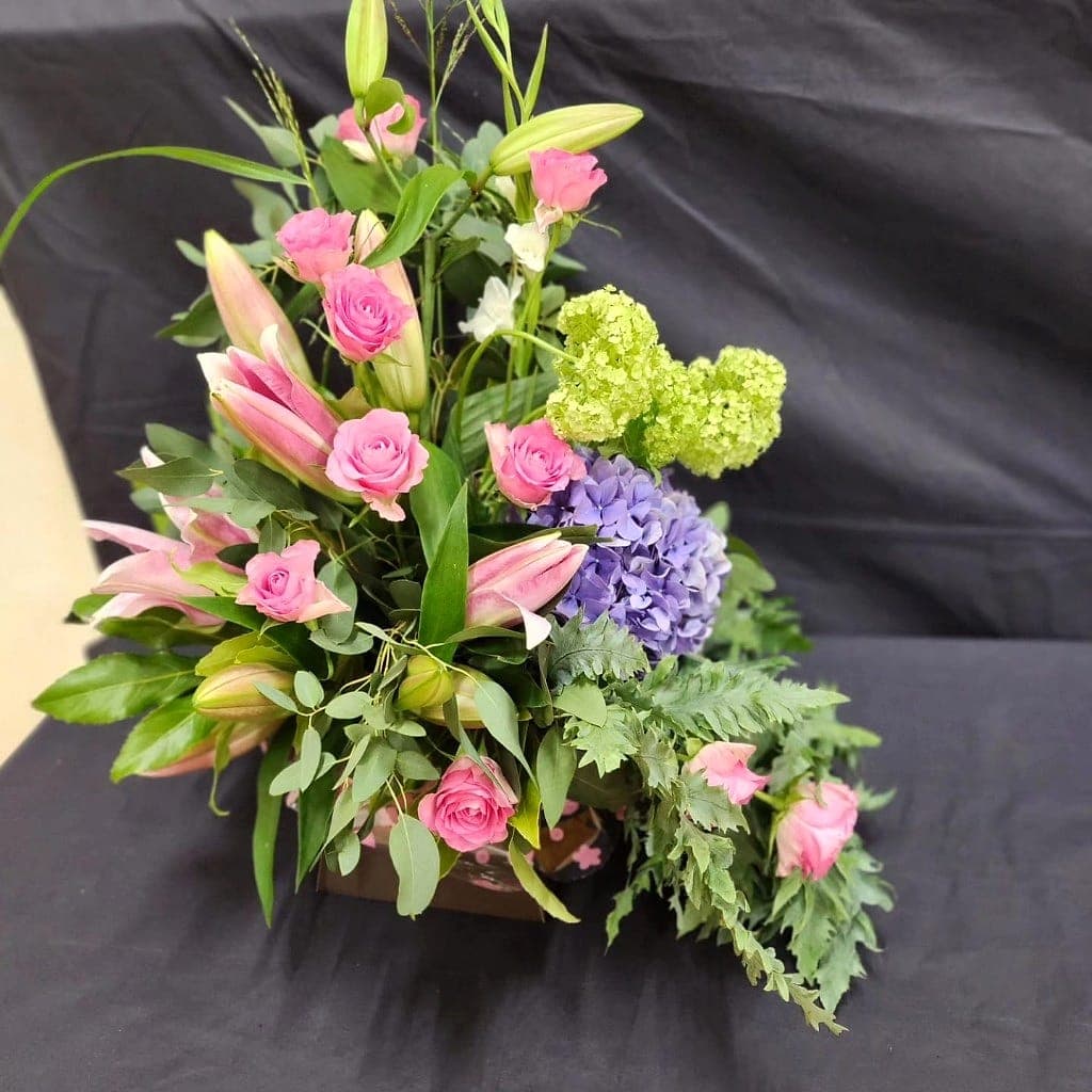Bespoke arrangement of pink roses, pink lilies and purple hydrangea with trailing greenery on a dark ground