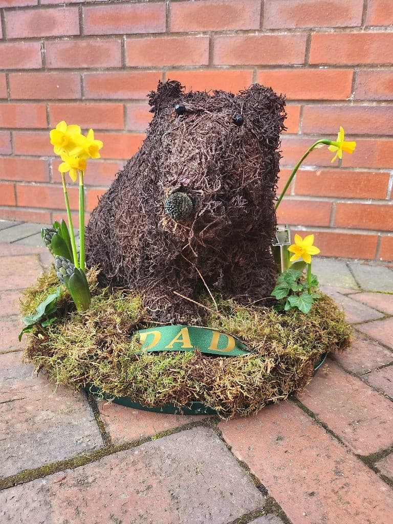 Moss-covered sitting bear tribute with yellow daffodils and a green ribbon reading DAD