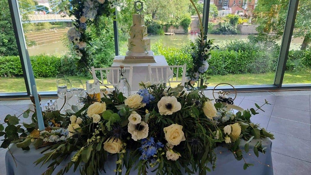 Wedding head table with a low white, cream and blue floral spray, taper candles and a gold hoop behind the cake